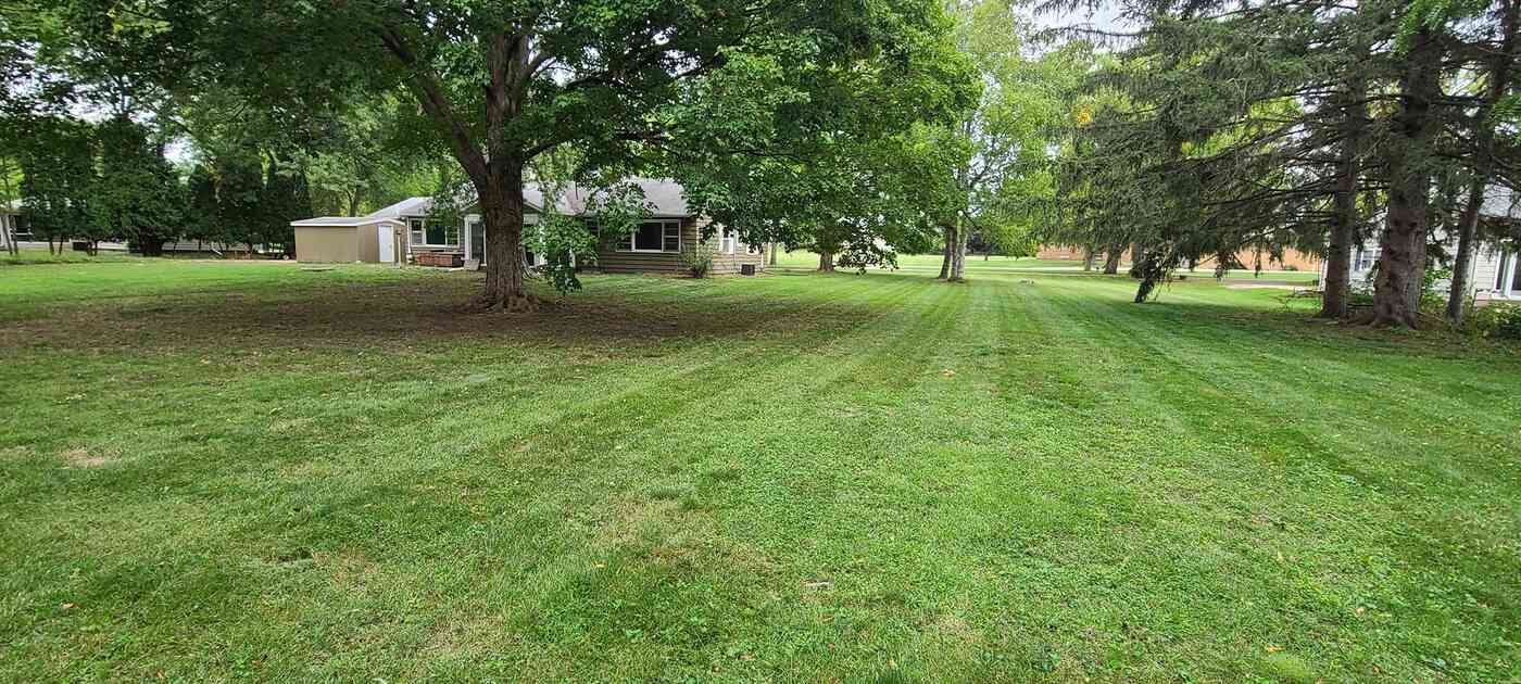 Large residential lawn mowed in even stripes under mature shade trees, with a ranch home in the background