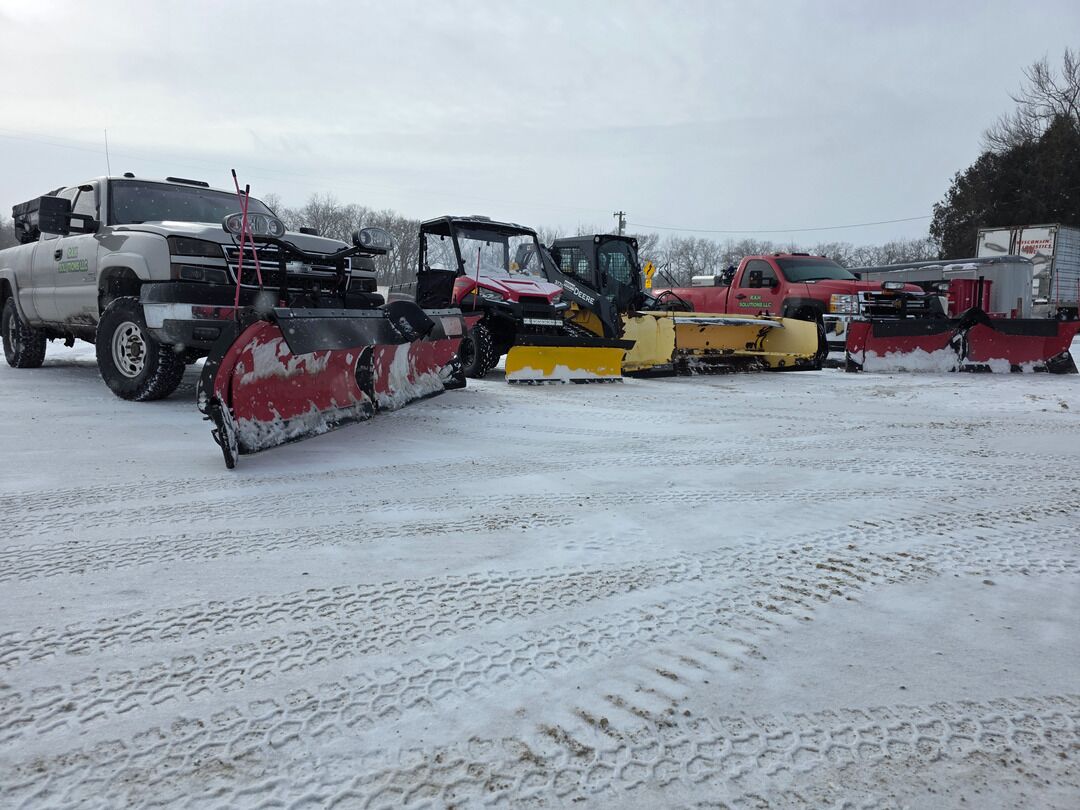 Plow trucks, a utility vehicle and a skid steer with snow blades lined up on a snowy lot